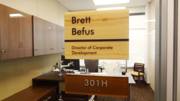 Office nameplate on glass door reads "Brett Befus, Director of Corporate Development" with room number "301H" below. Inside, there are desks, chairs, and office supplies.