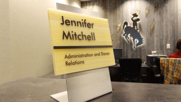 A desktop nameplate reads "Jennifer Mitchell, Administration and Donor Relations." The background shows a blurred office space with a person sitting at a desk and a wall decoration featuring a silhouette of a cowboy on a bucking horse.