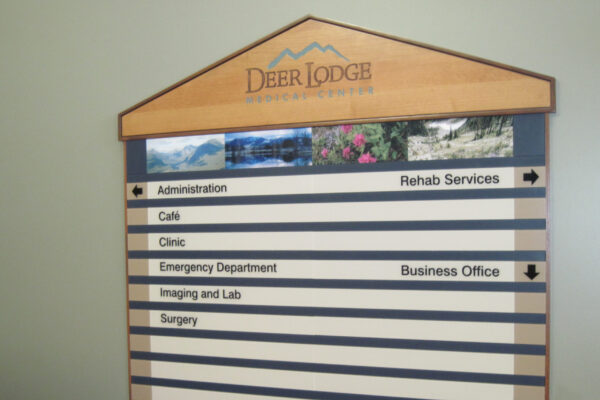 A wooden directory sign mounted on a wall, displaying various departments at Deer Lodge. Categories listed include Administration, Calls, Clinic, Emergency Department, Imaging and Lab, Surgery, Rehab Services, and Business Office, with arrows indicating directions.