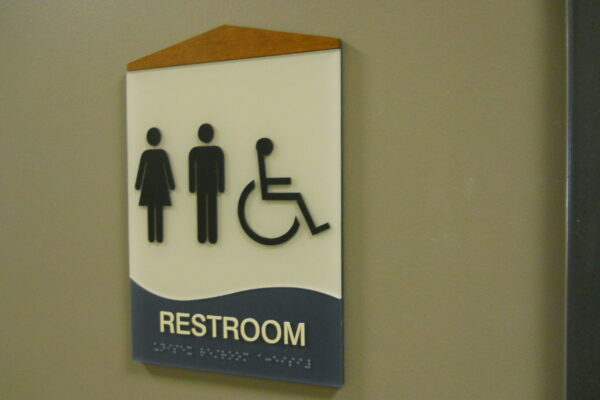 A restroom sign on a wall features symbols for women, men, and wheelchair accessibility above the word "Restroom." The top of the sign has a brown triangular accent.