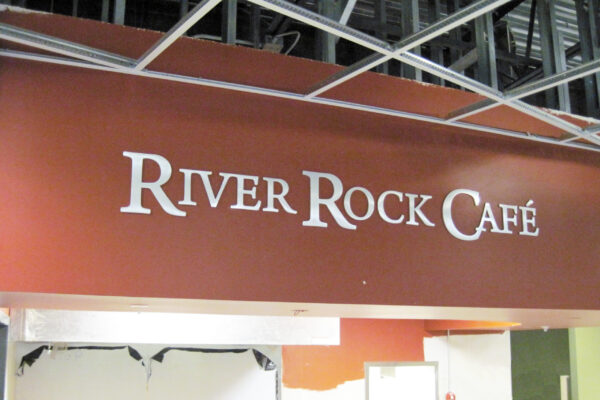 A sign for "River Rock Café" is displayed on a red-brown wall with white letters. The surrounding area appears to be under construction, with exposed ceiling beams and an unfinished section below the sign.