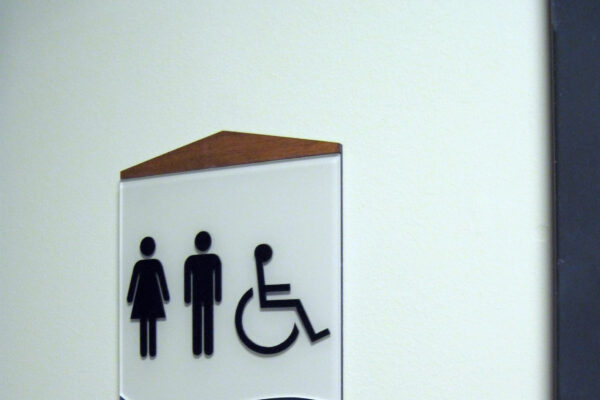 A restroom sign on a wall features symbols for women, men, and wheelchair accessibility above the word "Restroom." The top of the sign has a brown triangular accent.