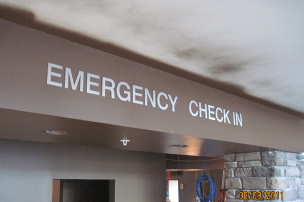 Signage inside a building reads "EMERGENCY CHECK IN" in bold white letters on a dark brown beam. The date on the bottom right corner shows 08/19/2011. The area underneath appears to be part of a reception or waiting area, with a stone wall visible in the background.