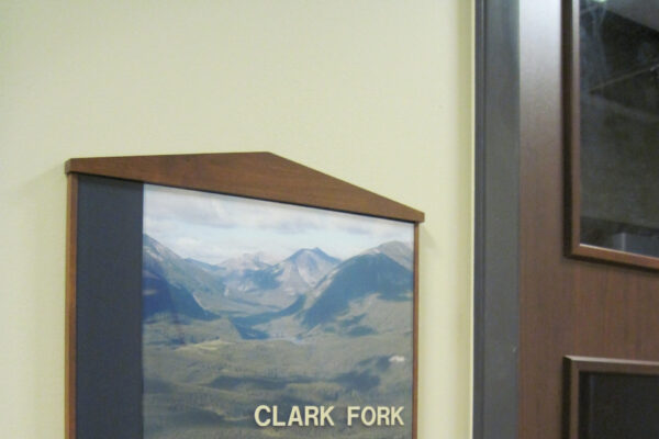A sign on a wall next to a door reads "Clark Fork" with a landscape image of mountains and a valley above the text. Below the nameplate, a smaller label indicates the room is "Available." The door is wooden with a metal handle.
