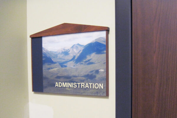 A wall sign for an administration office features a photograph of mountainous scenery beneath a wooden frame with a peak at the top. The word "ADMINISTRATION" is written in capital letters at the bottom of the image. A wooden door is partially visible beside the sign.
