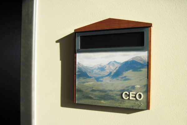 A sign mounted on a wall with a triangular wooden top and a rectangular frame. The sign has an image of a mountainous landscape with green valleys and a blue sky. The letters "CEO" are prominently displayed on the bottom right corner of the frame.