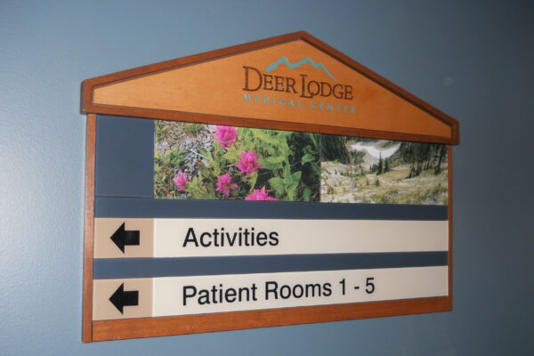 A wall-mounted directional sign with a wooden frame, labeled "Deer Lodge" at the top. Below are arrows pointing left for "Activities" and right for "Patient Rooms 1-5." The sign features small images of flowers and a nature scene.