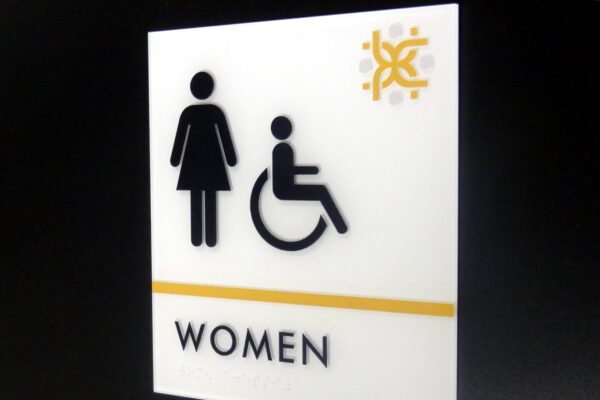 A sign with symbols indicating a women's restroom. It includes a pictogram of a woman and a wheelchair user. The word "WOMEN" is written in black text below the symbols, with a yellow line above the text. The upper right corner features a decorative logo.