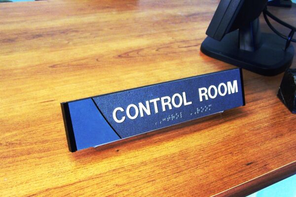 A small rectangular sign on a wooden surface reads "CONTROL ROOM" in bold white letters with Braille text below. The sign has a sleek black and blue color scheme, and a computer monitor is visible in the background.