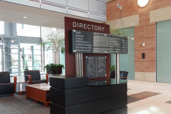 A directory sign inside a modern office building lists multiple companies and their suite numbers, including AMD, Broadcom, Cerience, CHD Bioscience, Crawford Technologies, First American Title, and Kaiser Permanente. The sign is made of glass and wood.
