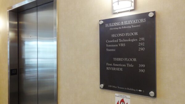 A directory board is mounted on a beige wall next to an elevator. It lists the names and room numbers of businesses on the second and third floors, such as Certified Technologies in room 201 and First American Title in room 300.