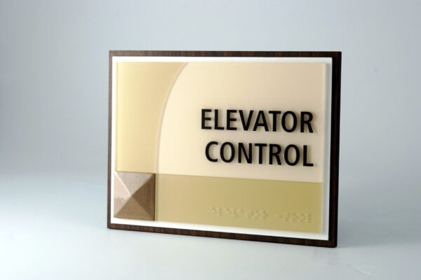 A sign on a light-colored wall reads "ELEVATOR CONTROL" in capital black letters. The sign features a geometric design with a textured square in the bottom left corner and curved lines in the background.
