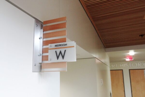 A wooden restroom sign with a large "W" is mounted on a wall. The wall and ceiling are beige, and a corridor with wooden doors is visible in the background. An exit sign is illuminated in red in the distance.