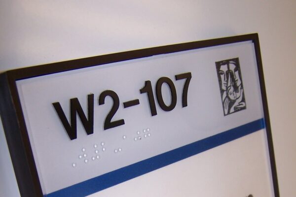 A room sign with the text "W2-107" and "Susan Berylian Client Services" is mounted on a wall. The sign has a blue stripe and a small icon in the top right corner. Braille text is above the blue stripe.