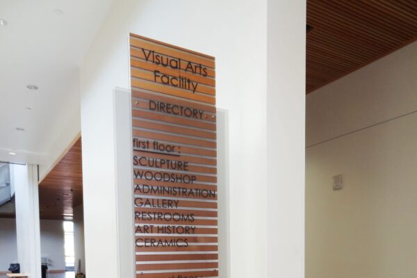 A tall, sleek directory sign inside a modern building, labeled "Visual Arts Facility." The list includes several art departments such as sculpture, painting, graphic design, and more. The grey and wood-accented sign stands on a polished concrete floor near a wooden table.