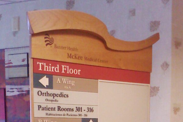 A stand-up directory sign in an indoor setting indicates locations within McKee Medical Center. The sign lists various departments on the third floor, including Orthopedics, Transitional Care, and Pulmonary. The background features soft-colored wallpaper.