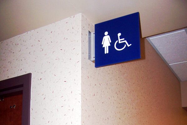 A corner in a building hallway shows a wall-mounted sign indicating restrooms for women and those with disabilities. Below, another similar sign is positioned on the wall next to the restroom door. Both signs feature accessible icons.