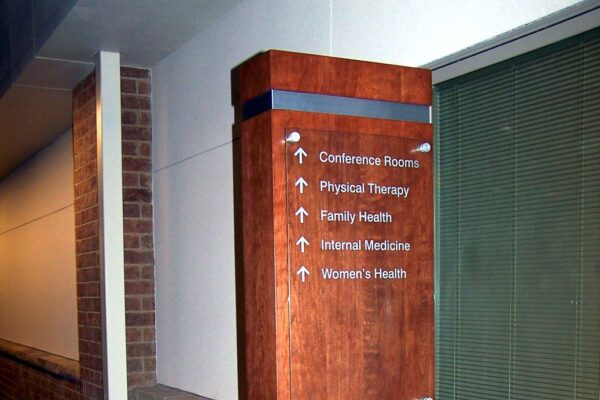 A wooden directional signpost in a hallway points to different locations including conference rooms, physical therapy, family health, internal medicine, and women's health. The sign features white text and arrows on a wood panel set against a brick wall and carpeted floor.