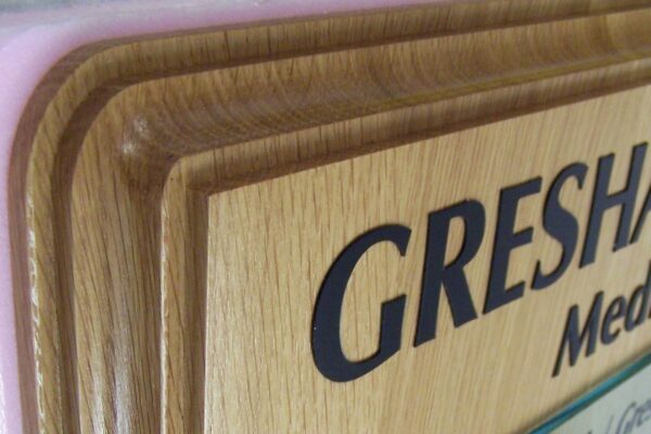 A close-up shot of a wooden directory sign with glass panels on a wall. It has several sections listing different names and titles. The partial text "GRESHAM" is visible at the top. The layers of the sign create a three-dimensional effect.