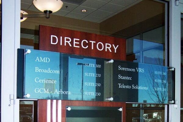 A directory sign mounted on a wooden board in a lobby area. The board lists various company names such as AMD, Broadcom, and others, with corresponding suite numbers. Below the listings is a blank black space for additional information. Glass doors are visible in the background.