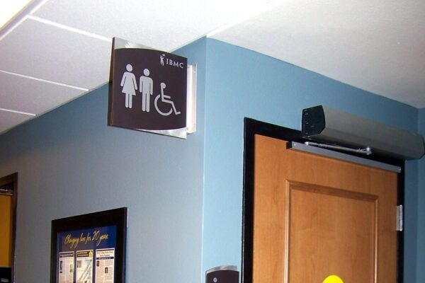 A restroom door with a wooden finish is shown. Above the door is a sign indicating it is a unisex and accessible restroom. A wall-mounted sign adjacent to the door repeats the same information. A yellow caution sticker is on the door, which is closed.