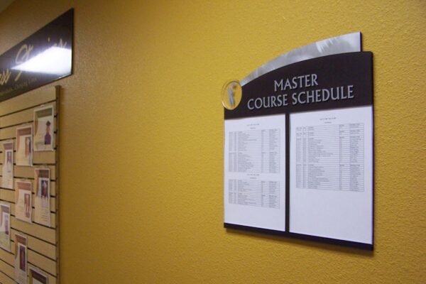 A wall-mounted display titled "Master Course Schedule" on a yellow wall. The schedule consists of two white sheets with text. Nearby on the left, a bulletin board contains various posted items, including photos and papers. The scene is well-lit.