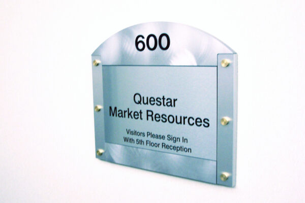 A metallic office door sign features the text "600 Questar Market Resources" with a note below reading "Visitors Please Sign In With 5th Floor Reception." The sign has a silver background with an arched top, mounted on a white wall.