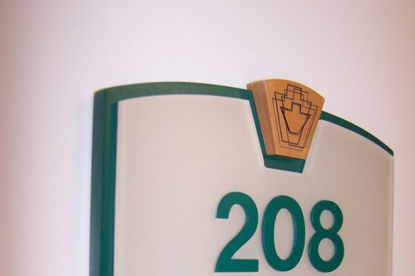 A wall-mounted plaque with the number 208 in green, featuring an emblem at the top. Below is a sliding nameplate showing "MR & Mrs. James N. Gleason" with the option to slide in text. The sign is accented with green borders.