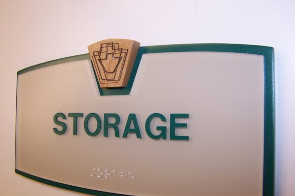 A frosted glass sign with green borders and a wooden emblem at the top, displaying the word "STORAGE" in green uppercase letters. Below the text, there is braille writing. The sign is mounted on a white wall.