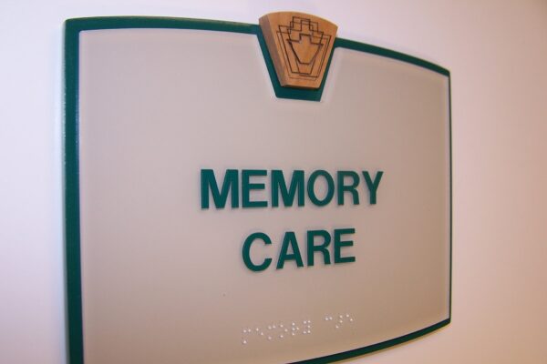 A frosted glass sign with green borders and a wooden emblem at the top, displaying the words "MEMORY CARE" in green uppercase letters. Below the text, there is braille writing. The sign is mounted on a white wall.