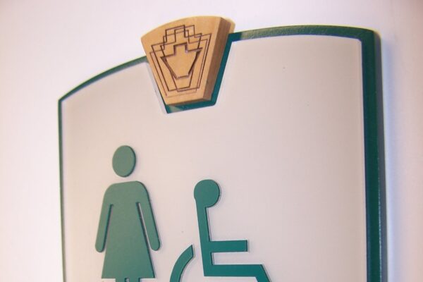 A restroom sign for women with a green border. The sign features symbols for a woman and a person in a wheelchair, indicating the restroom is accessible. The word "WOMEN" is written below the symbols in green text. Braille is included beneath the text.