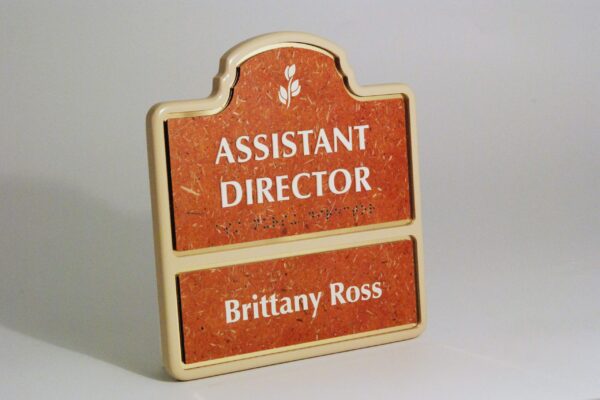 A sign with a red background and a beige border. The top part reads "ASSISTANT DIRECTOR" in white text, while the bottom part displays the name "Brittany Ross" in the same white text.