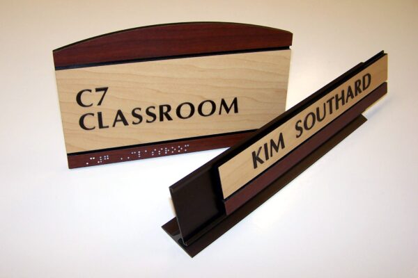 A classroom sign labeled "C7 Classroom" with braille text beneath it is displayed beside a desk nameplate that reads "Kim Southard." Both signs feature a combination of light wood and dark accents, and are positioned on a white background.