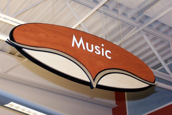 A hanging brown and white oval sign in a store or library, displaying the word "Music" in white text. The ceiling structure and lights are partially visible in the background.