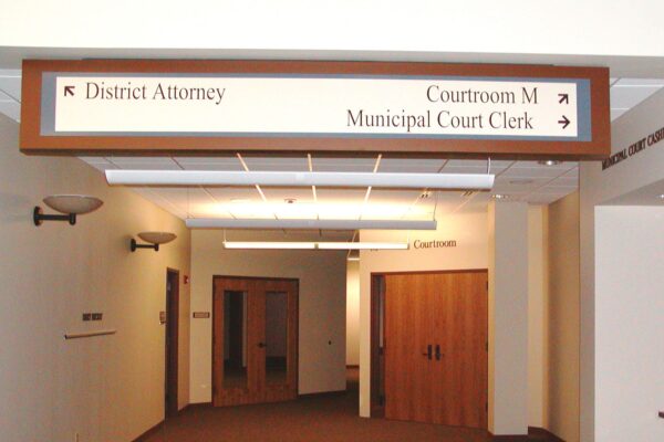 A hallway in a building with signs on the ceiling directing to the offices of the District Attorney, Municipal Court Clerk, and Courtroom M. The walls are cream-colored and several doors are visible along the corridor. Wall sconces provide lighting.