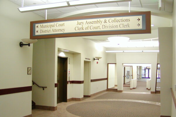 A hallway inside a courthouse with a signpost hanging from the ceiling. The sign directs to Municipal Court, District Attorney, Jury Assembly & Collections, and Clerk of Court, Division Clerk. The corridor features beige walls and carpeted floors.