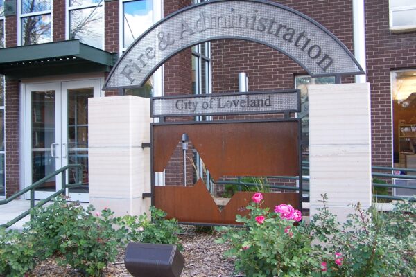 A decorative metal sign at the entrance of a brick building reads "Fire & Administration, City of Loveland." The bottom portion of the sign features a m cut-out design. There are flowering bushes and greenery in front of the sign.