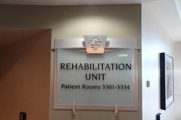 A sign mounted on a light-colored wall indicates the location of a Rehabilitation Unit in a medical facility. The sign includes room numbers for patient rooms, specifically 5301 to 5334. The design features a medical emblem above the text.