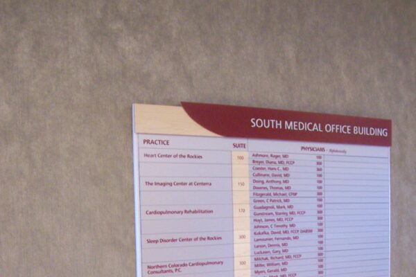 Directory board on a gray wall in the South Medical Office Building. The board lists various office numbers, doctor names, and specialties. A small portion of a cushioned chair is visible at the bottom left corner of the image.