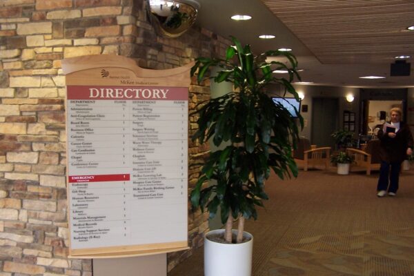 A directory sign is displayed in a hallway with a stone wall on the left. The sign lists different locations within the building. Adjacent to the sign is a large potted plant. The hallway leads to more rooms in the background, with a few people visible.