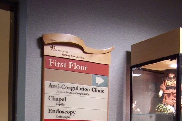 A directory sign on a wall lists locations on the first floor of a building, including the Anti-Coagulation Clinic, Chapel, Endoscopy, Emergency, Employee Health, Gift Shop, Radiology (X-Ray), Patient Registration, Warm Water Therapy, and McKee Breast Center.