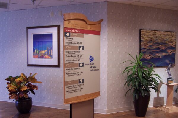 A hospital directory sign stands in a hallway, listing departments on the second floor. Two framed pictures hang on the wall behind it. Potted plants are positioned nearby, and the hallway has a wooden floor. .