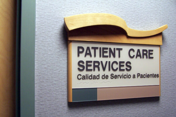 A wall-mounted sign reads "PATIENT CARE SERVICES" with the Spanish translation "Calidad de Servicio a Pacientes" below it. The sign is bordered by a light wooden top section and colored panels at the bottom. The background wall is light gray.