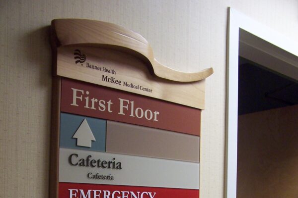 A wall-mounted directory sign inside a building, indicating locations on the first floor. It lists the cafeteria, emergency, lobby/information, board room, and McKee Surgery Center lobby with arrows pointing the directions to each area.