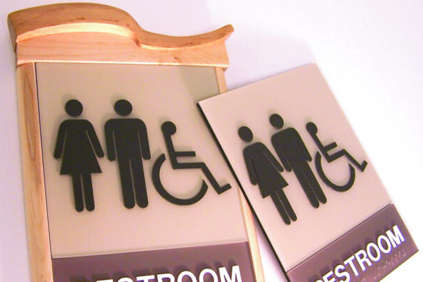 A restroom sign with icons representing male, female, and accessible facilities. The sign consists of a main wooden plaque with a smaller detachable portion displaying the same icons. Below the icons, the word "Restroom" is written in bold letters.