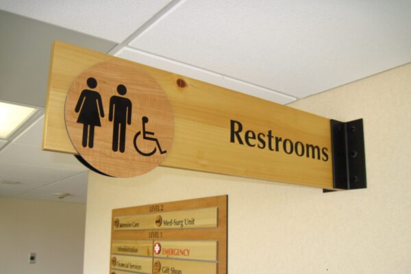 A wooden sign with a female and male restroom icon, a wheelchair accessibility symbol, and the word "Restrooms," is mounted on a beige wall. Below the sign is a directory listing various locations within the building.