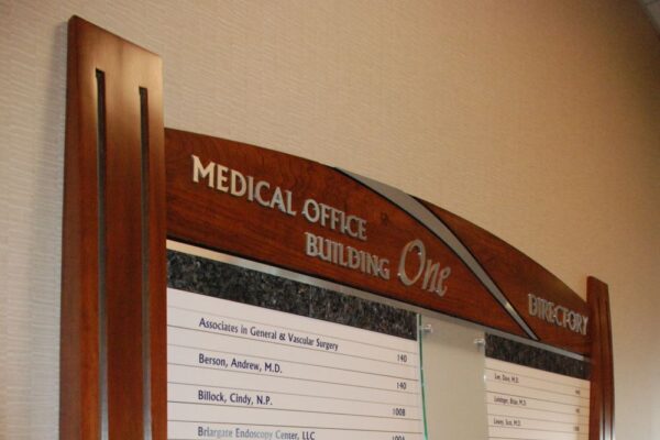 A directory sign inside a building labeled "Medical Office Building One" lists names and room numbers of various doctors. The sign has a wooden frame with a curved top and features a partial glass panel in the center.