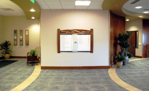 The image shows a spacious, well-lit interior of a building lobby with tiled flooring and neutral-colored walls. A directory board is mounted on the central wall, flanked by potted plants. A hallway is present on the right side, leading to other rooms or areas.