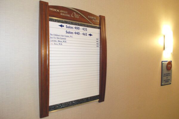 A mounted board with a wooden frame displays a list of events scheduled for the day. To the right, a wall-mounted directory sign can be seen. The board is titled "MEDICAL OFFICE" and indicates rooms and times for appointments. The setting appears to be in a corridor.