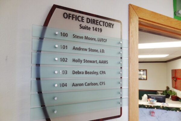 A glass office directory sign mounted on a beige wall lists suite 1419 occupants: 100 Steve Moore, LUTCF; 101 Andrew Stone, J.D.; 102 Holly Stewart, AAMS; 103 Debra Beasley, CPN; 104 Aaron Carlson, CFS. The entrance to a well-lit office space is visible to the right.